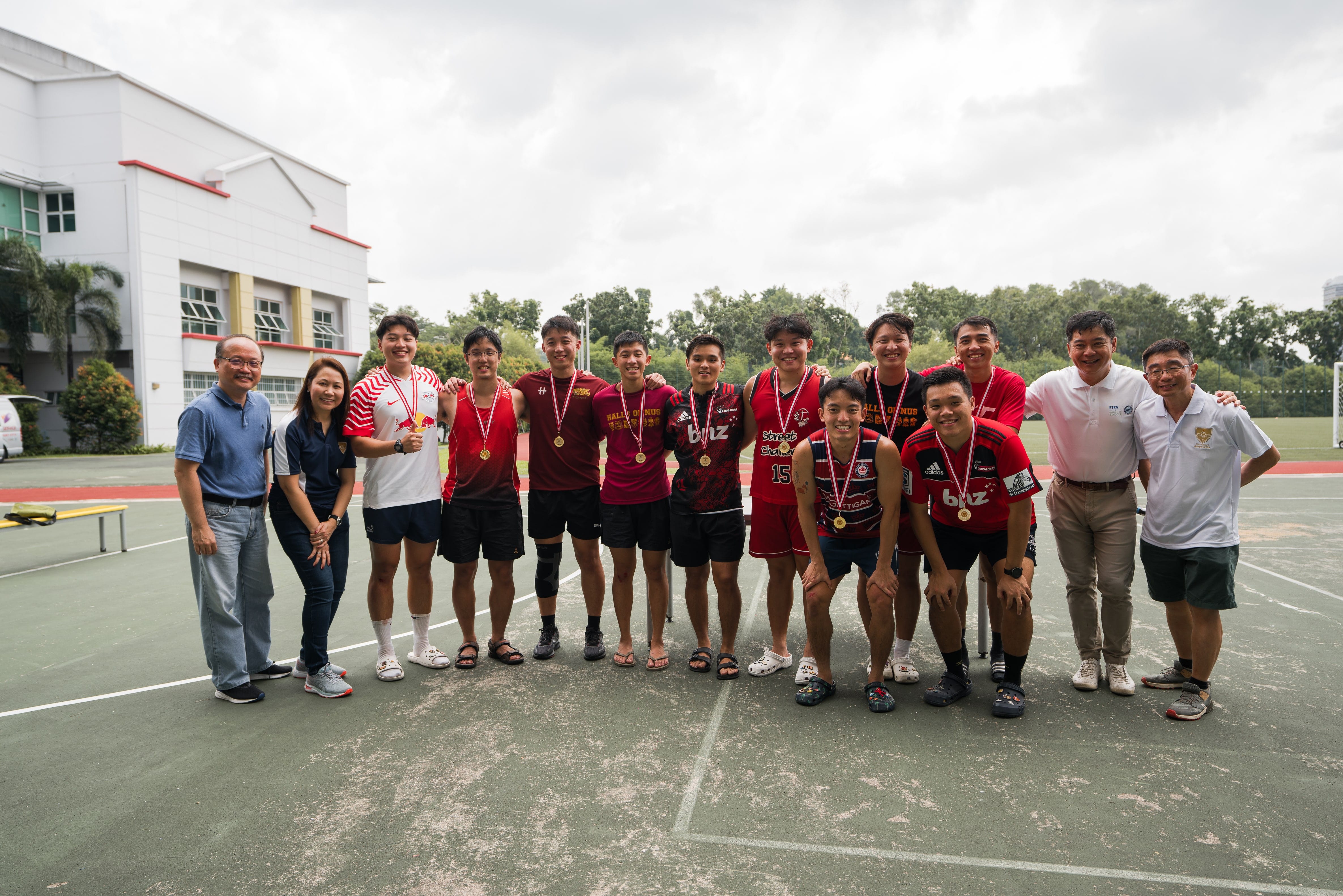 A group of young athletes wearing gold medals pose together on an outdoor sports court near a school building.
